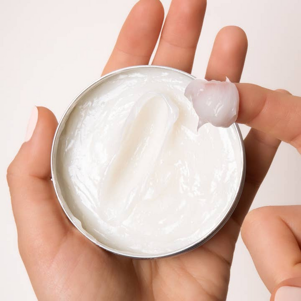 Hand holding a small container of white cream with a visible scoop on a light background