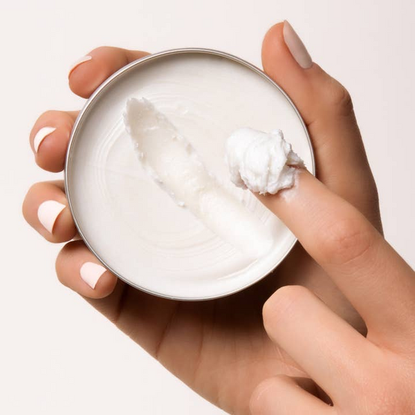 Hand holding a small container of white cream with a visible scoop on a light background