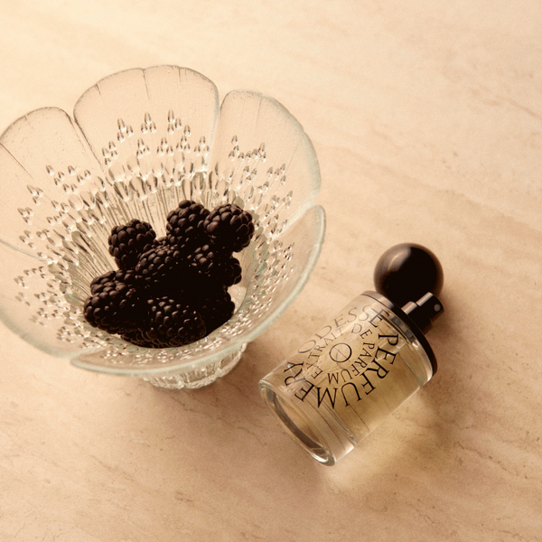 Small perfume bottle next to a glass dish with blackberries on a beige surface
