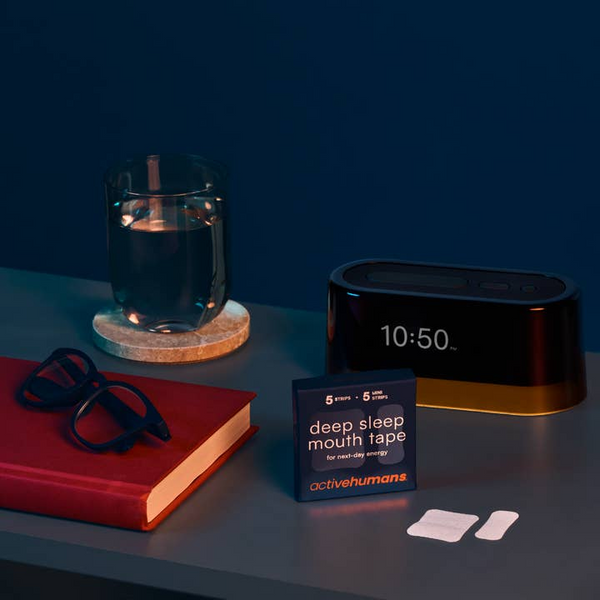 Tabletop scene with a clock, glass of water, book, glasses, and a box labeled 'deep sleep mouth tape' on a dark surface.