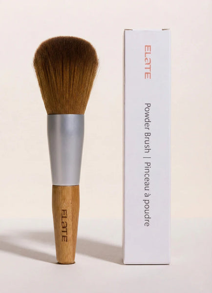 Elate powder brush with wooden handle and silver ferrule next to its packaging on a light beige background