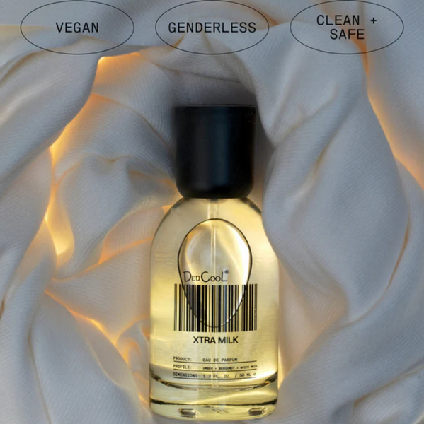 Perfume bottle labeled 'Xtra Milk' with 'Vegan', 'Genderless', and 'Clean + Safe' labels on a white fabric background.
