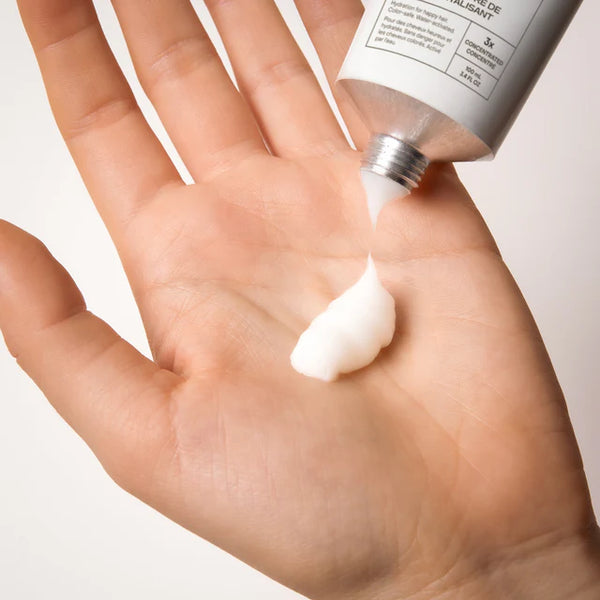 Hand applying cream from a tube of The Deep Conditioning Concentrate on a plain background
