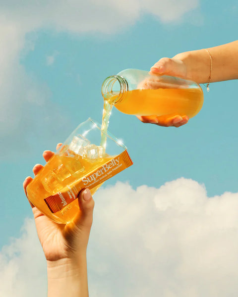 Person pouring orange Mango Passion Fruit SuperBelly by Blume from a bottle into another bottle labeled Blume 'SuperBelly' against a blue sky.
