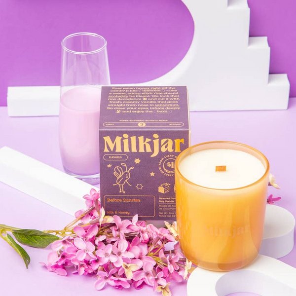 Milkjar candle with packaging, glass, and flowers on a purple background