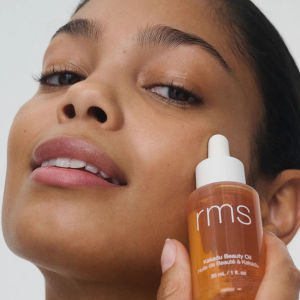Person holding a bottle of RMS Beauty oil near their face against a neutral background