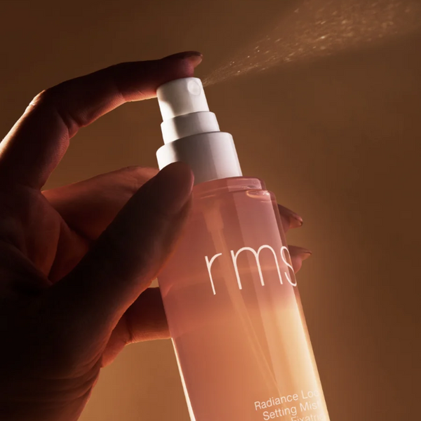 Hand holding a RMS beauty spray bottle against a warm brown background