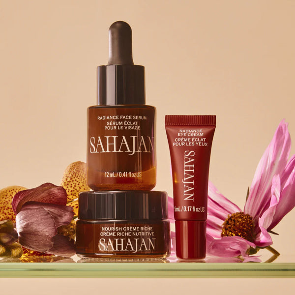 Sahajan skincare products including a serum, cream, and eye balm on a beige background with flowers.