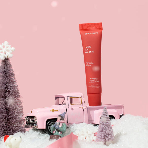Red skincare tube labeled 'ESW Beauty' with a pink toy truck and Christmas trees on a pink background