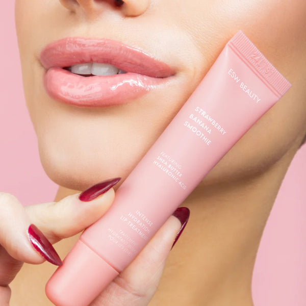 Pink lip balm tube labeled Strawberry Banana Smoothie Lip Treatment held by a hand with red nail polish against a pink background.