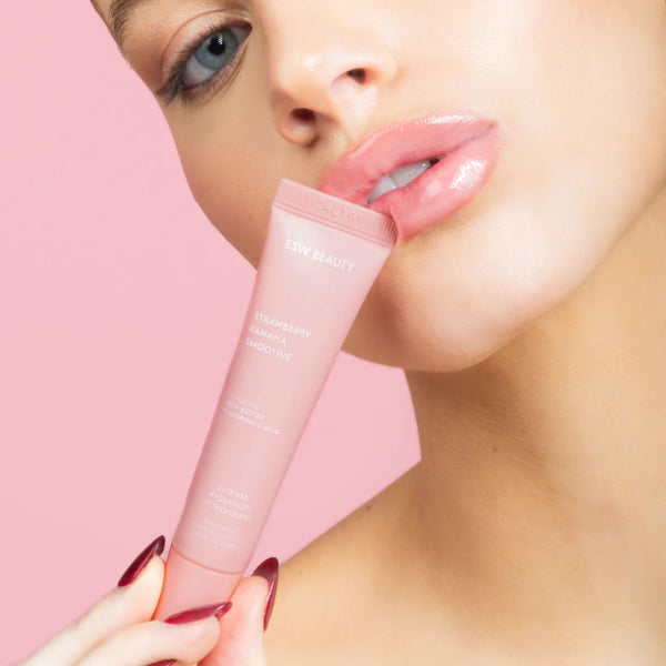 Woman holding a pink lip balm with a soft pink background wearing Strawberry Banana Smoothie Lip Treatment